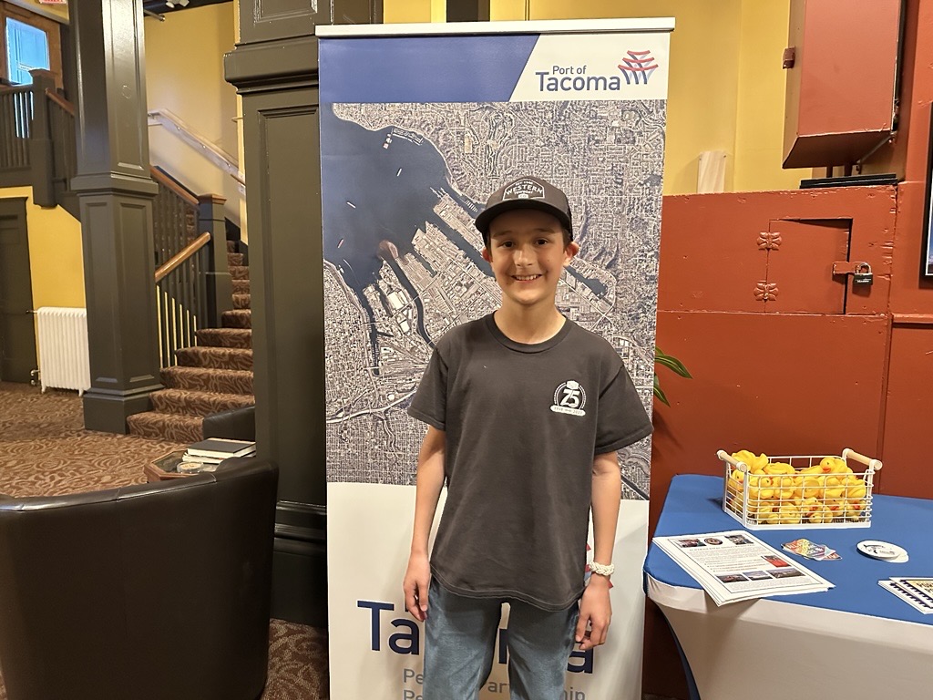 An adolescent boy stands in front of a Port of Tacoma map near a table.