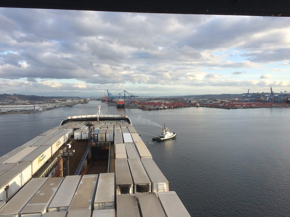 A boat with an opening in the middle sits atop a waterway with a tugboat in the background.