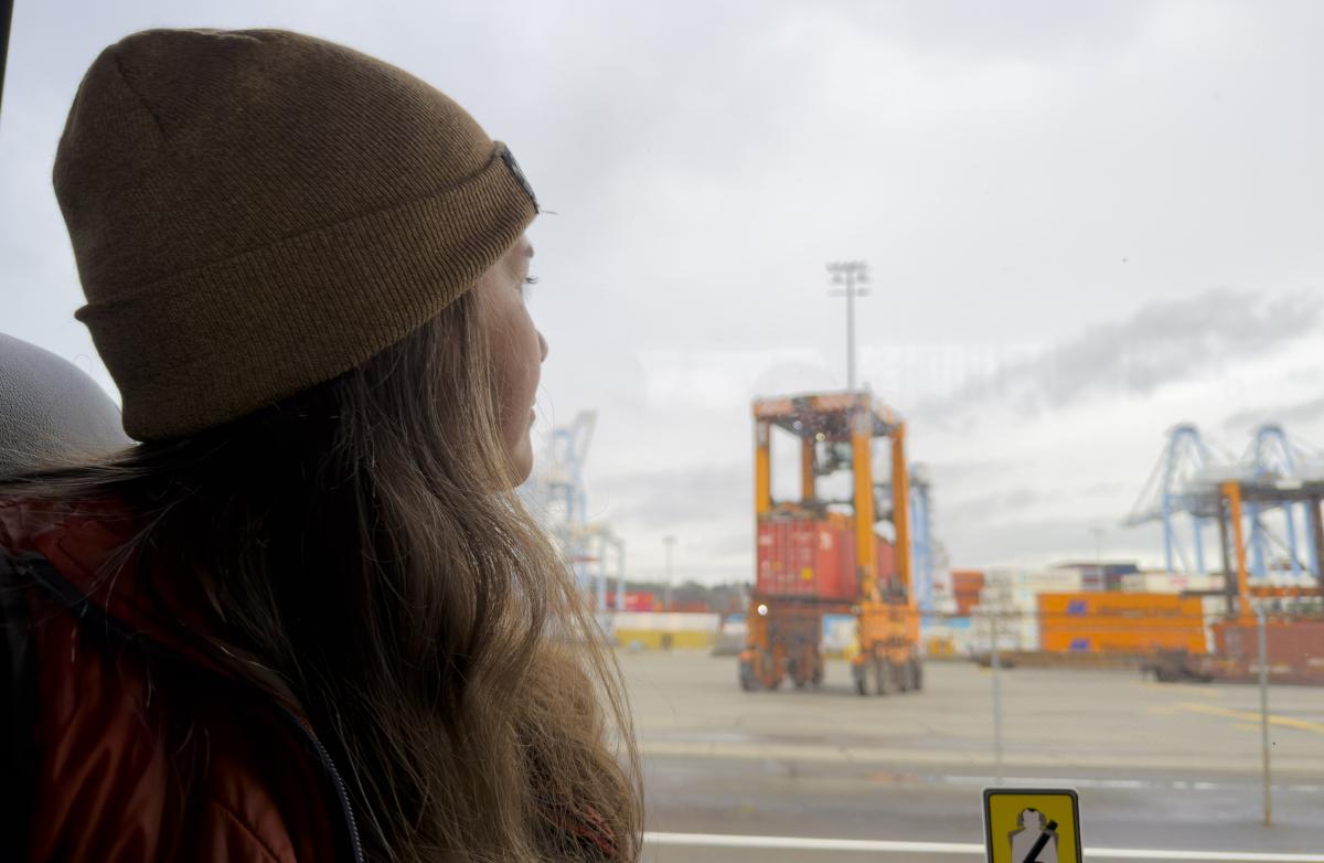 A woman with long hair wearing a beanie looks out a bus window at a port terminal