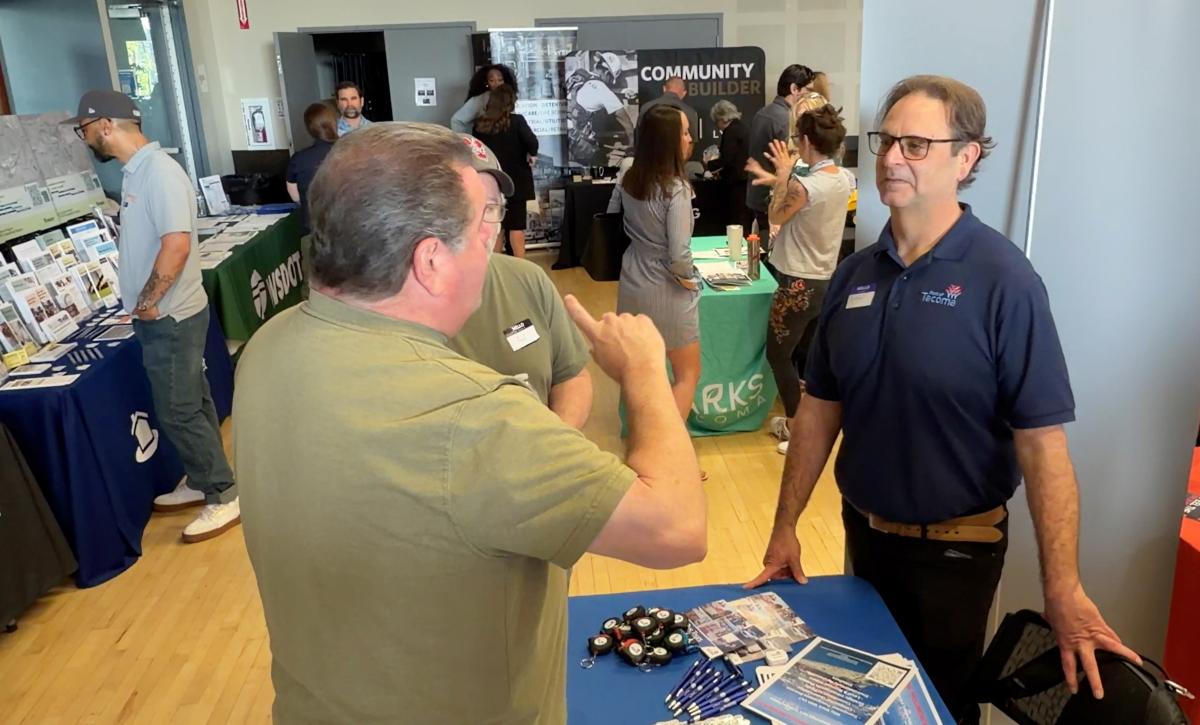 A man in a polo shirt behind a table speaks to two other men at a networking event.
