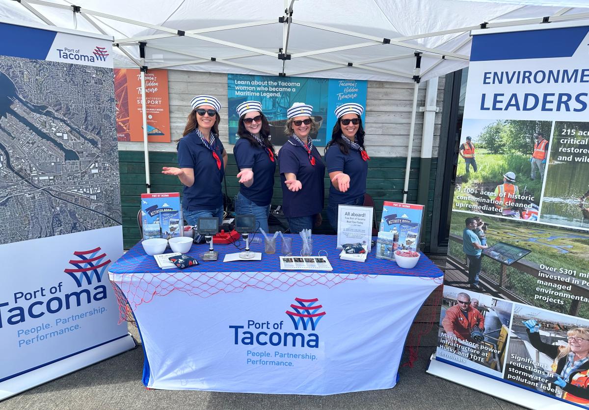 Four women in sailor hats hold their hands out while standing behind a Port booth at Oceanfest 2024.